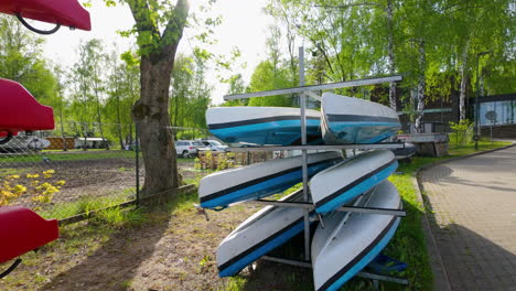 stack-of-blue-and-white-kayaks-stored-on-metal-racks-near-a-tree-and-a-parking-area,-with-red-kayaks-in-the-foreground