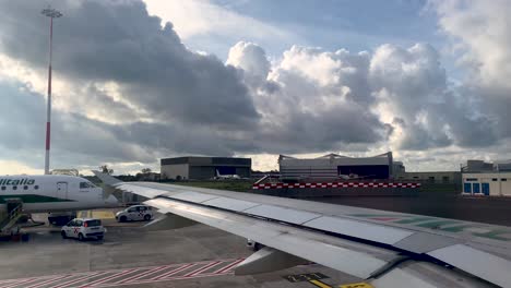 Vista-Desde-El-Interior-Del-Ala-Del-Avión-Mientras-El-Espectador-Pasa-Por-Delante-De-Los-Vehículos-De-Alitalia-En-Tierra-En-El-Aeropuerto-Internacional-De-Roma.