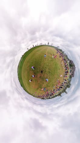 360-degree-view-of-an-egg-hunting-event,-capturing-families-and-children-scattered-across-a-grassy-field-under-a-cloudy-sky