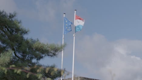 Two-flags,-one-of-the-European-Union-and-the-other-of-Luxembourg,-wave-proudly-against-a-blue-sky-with-soft-clouds