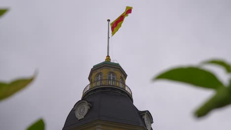 Una-Bandera-Ondeando-Con-Fuerte-Viento-Desde-Una-Torre-De-Castillo-Con-Un-Cielo-Gris-Y-Hojas-Desenfocadas