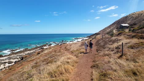 Una-Pareja-Caminando-Por-Un-Remoto-Camino-De-Tierra-Con-Una-Pequeña-Choza-A-Su-Derecha-Y-El-Océano-Azul-Brillante-Y-La-Playa-De-Arena-Blanca-A-Su-Izquierda.