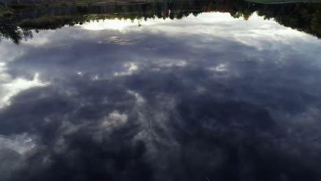 Slow-moving-aerial-drone-shot-over-a-calm-pond-with-reflections-of-the-clouds-in-the-sky