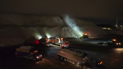 An-establishing-drone-shot-of-large-trucks-dumping-trailers-full-of-snow-and-ice-at-a-snowbank,-a-large-bulldozer-sprays-snow-up-high-into-the-air-building-the-bank-at-night