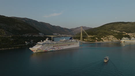 Aerial-5K-Drone-Over-Dubrovnik-Harbor-With-Norwegian-Cruise-Ship-Sailing-Beyond-Cable-Bridge-In-Croatia