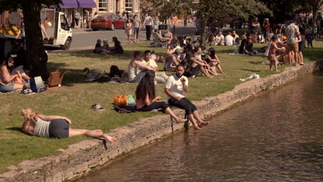 Durante-La-Pandemia-De-Covid-19,-Se-Ve-A-La-Gente-Disfrutando-De-Un-Día-Soleado-Al-Lado-De-Un-Canal-En-Bourton-on-the-water,-Cotswold,-Reino-Unido