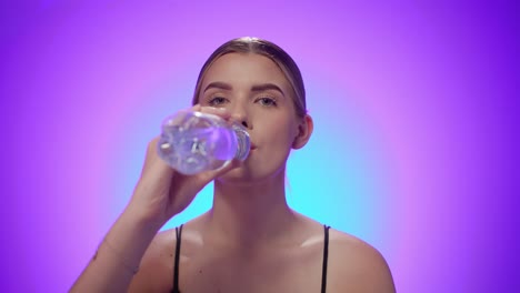 Durstige-Frau-Trinkt-Wasser-Aus-Einer-Transparenten-Plastikflasche,-Violetter-Hintergrund