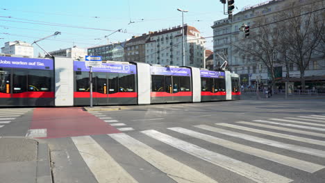 Eine-Handaufnahme-Einer-Kreuzung-In-Wien-Mit-Zwei-Vorbeifahrenden-Straßenbahnen-Und-Menschen,-Die-Im-Hintergrund-über-Einen-Zebrastreifen-Gehen