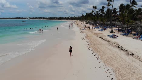 Frau,-Die-An-Einem-Wunderschönen-Tropischen-Strand-Mit-Palmen-In-Der-Dominikanischen-Republik-Spazieren-Geht,-Luftaufnahme-Mit-Absteigender-Neigung-Nach-Oben