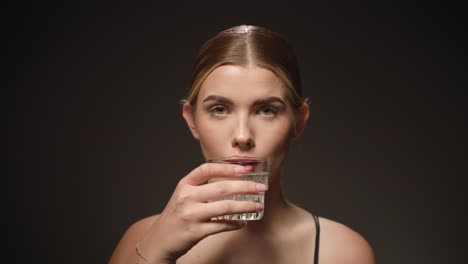 Una-Mujer-Seria-Toma-Un-Sorbo-De-Un-Vaso-De-Agua-Transparente,-Fondo-Negro