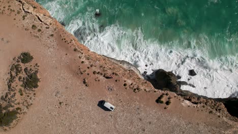 Birdseye-view-of-a-couple-standing-near-a-camper-van-on-a-cliff-edge-with-the-waves-crashing-against-the-rocks-below