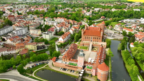 Una-Vista-Aérea-Que-Destaca-El-Castillo-Histórico-Y-Los-Edificios-Circundantes-En-Lidzbark-Warmiński,-Con-Un-Río-Que-Atraviesa-La-Escena-Y-La-Ciudad-Al-Fondo.