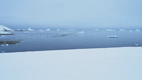 Verschneite-Winterlandschaft-Mit-Eisbergen-In-Der-Antarktis,-Schnee--Und-Eisberglandschaft-Auf-Der-Antarktischen-Halbinsel-Mit-Blick-Auf-Das-Meerwasser-Vom-Antarktischen-Festland-Aus