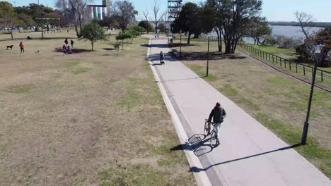 Mann-Läuft-Neben-Seinem-Fahrrad-Am-Flussufer-Neben-Einem-Park,-In-Dem-Die-Leute-Spaß-Haben