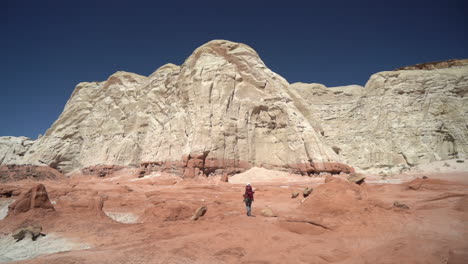 Lonely-Woman-With-Backpack-Walking-in-Dry-Desert-Landscape-Under-Sandstone-Hills-on-Hot-Sunny-Day,-Back-View