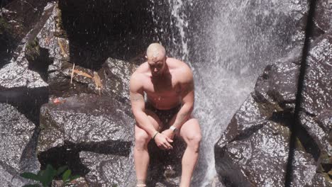 A-muscular-man-sits-under-the-cascading-waters-of-Kanto-Lampo-Waterfall-in-Bali,-Indonesia