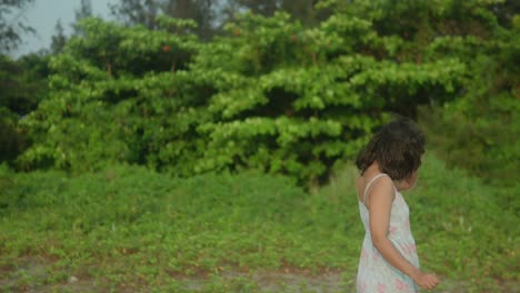 Young-woman-in-a-floral-dress-walks-away-on-a-sandy-path,-lush-greenery-in-the-background