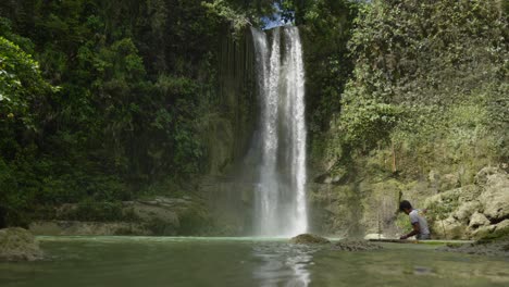 At-Camugao-Falls-in-the-Philippines,-water-cascades-downward-as-a-local-craftsman-diligently-works-on-his-surfboard,-a-vivid-illustration-of-travel-and-exploration