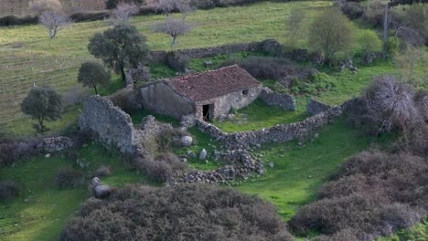 Drone-flight-in-which-we-visualize-an-old-stone-house,-probably-for-agricultural-use,-surrounded-by-abandoned-crop-fields,-with-almost-the-entire-roof-preserved-and-has-a-patio-with-a-wall-in-Madrid