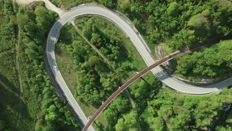 Static-aerial-overhead-view-of-a-winding-road-intersected-by-an-old-railway-bridge-in-the-middle-of-a-forest