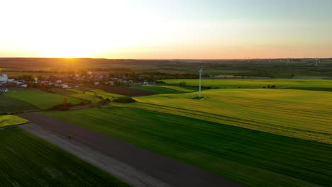 Aerial-view-of-a-village-surrounded-by-green-fields-at-sunset-with-wind-turbines-in-the-background