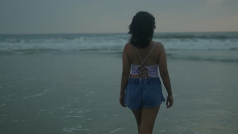 Mujer-Joven-Con-Pantalones-Cortos-Azules-Parada-Contemplativamente-Junto-Al-Océano-Al-Atardecer,-Suaves-Olas-Rompiendo-En-La-Orilla