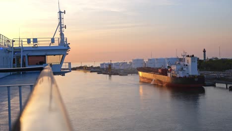 Im-Hafengebiet-Bewegt-Sich-Eine-RoRo-Fähre-Langsam-Auf-Ein-Rot-schwarzes-Frachtschiff-Zu,-Das-In-Einem-Wunderschönen-Abendsonnenuntergang-Vor-Anker-Liegt.
