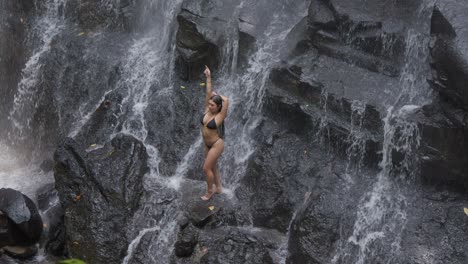 A-woman-in-a-black-bikini-is-posing-on-the-rocks-under-the-cascading-water-at-Kanto-Lampo-Waterfall-in-Bali