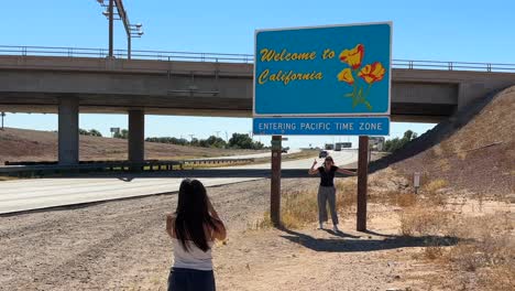 Young-female-tourists-taking-pictures-with-Welcome-to-California-state-sign