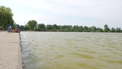 Elderly-woman-in-distance-wearing-bikini-sitting-on-cement-edge-of-city-park-lake