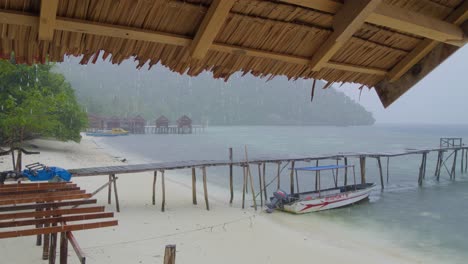 the-tropical-rain-shower-on-Kri-Island-in-the-Raja-Ampat-Archipelago,-as-it-cascades-onto-the-sea-and-sandy-beach,-seen-from-the-shelter-of-wooden-cottage