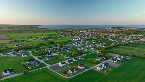 Aerial-view-of-a-village-in-Puck,-Połczyno,-with-numerous-houses-surrounded-by-green-fields-and-a-distant-body-of-water-at-sunset