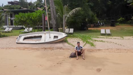 Male-Sitting-On-Ao-Tan-Beach-Beside-Empty-Rustic-Boat