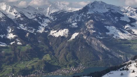 Vista-Aérea-De-Las-Montañas-Cubiertas-De-Nieve-Con-Exuberante-Y-Lago-En-Glarus,-Suiza