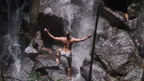 Un-Hombre-Posa-Y-Flexiona-Sus-Músculos-Bajo-El-Agua-De-La-Cascada-Kanto-Lampo-En-Bali
