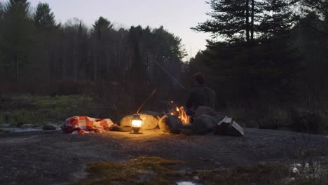 Man-fishing-by-a-campfire-with-a-lantern-and-blanket-in-a-forest-at-dusk,-surrounded-by-trees-and-nature