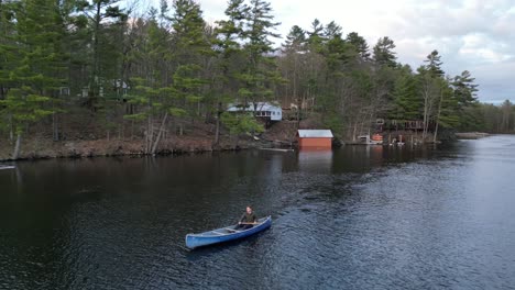 Person-paddling-a-canoe-on-a-forest-river-with-cabins-along-the-banks-at-dawn,-surrounded-by-nature-and-calm-water