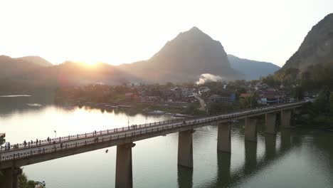 drone-shot-of-two-lane-bridge-at-sunset-in-the-mountain-town-of-Nong-Khiaw-in-Laos,-Southeast-Asia
