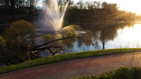 Dzirnavu-pond-with-fountain-with-magical-sun-rays,-side-flying-aerial-view
