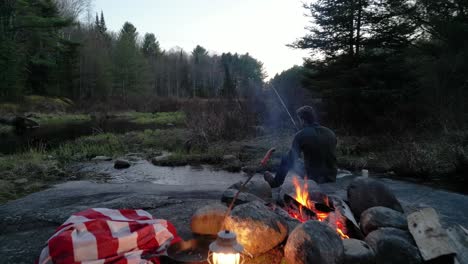 Man-fishing-by-a-campfire-with-a-lantern-and-blanket-in-a-forest-at-twilight,-surrounded-by-trees-and-the-tranquil-sounds-of-nature