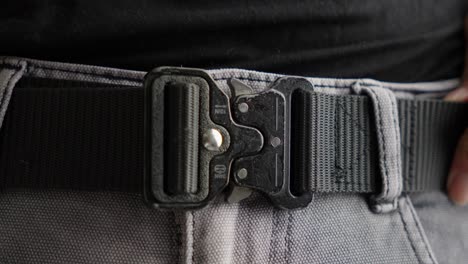 Fasten-And-Buckle-Black-Tactical-Belt-With-Hands-On-Gray-Jeans,-Close-Up