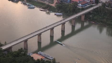 Barco-Pasando-Por-Debajo-De-Un-Puente-Sobre-El-Río-En-La-Ciudad-Montañosa-De-Nong-Khiaw-En-Laos,-Sudeste-De-Asia