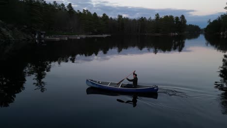Person-canoeing-on-a-serene-lake-at-twilight,-with-reflections-of-trees-on-the-calm-water-and-a-peaceful-atmosphere