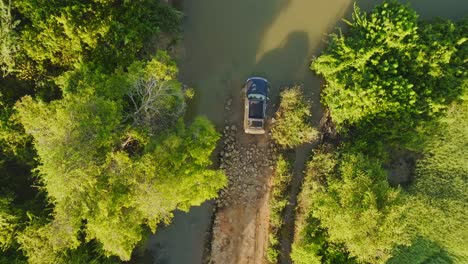 Luftaufnahme-Von-Oben-Nach-Unten-Auf-Einem-Schwarzen-Geländewagen-Auf-Einer-Grünen-Waldstraße,-Die-An-Einem-Sonnigen-Tag-In-Afrika-Durch-Eine-Tiefe-Schlammpfütze-Führt