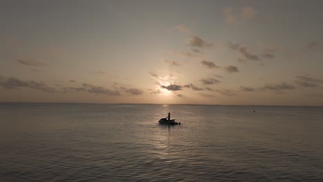 Persona-Del-Círculo-De-Drones-En-Silueta-De-Moto-Acuática-En-Una-Suave-Y-Suave-Puesta-De-Sol-Con-Horizonte-Oceánico