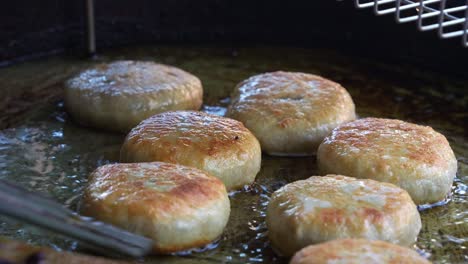 Close-up-shot-capturing-the-food-stall-vendor-uses-kitchen-tongs-to-flip-the-freshly-made-meat-pies,-sizzling-in-the-hot-oil-pan,-pan-frying-and-cooking-until-golden-brown,-a-popular-Asian-food