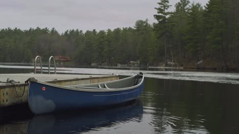 A-blue-canoe-is-docked-at-a-wooden-pier-on-a-calm-lake,-surrounded-by-tall-pine-trees-and-a-serene-natural-landscape,-under-an-overcast-sky