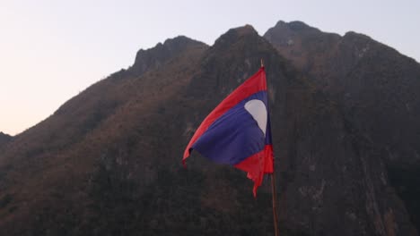 Bandera-De-Laos-Ondeando-Al-Viento-En-La-Ciudad-Montañosa-De-Nong-Khiaw-En-Laos,-Sudeste-Asiático