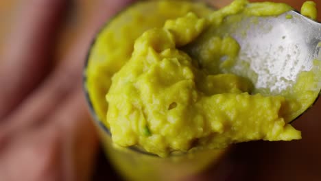 Scooping-Mixing-Yellow-Avocado-Puree-With-Spoon-Into-Glass-Pot,-Close-Up