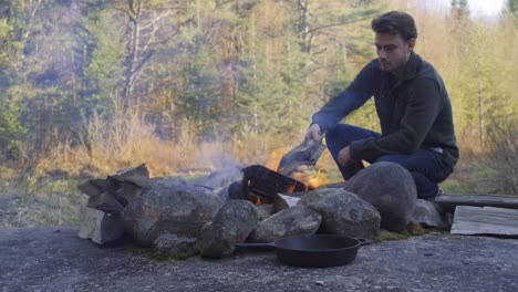 Un-Hombre-Cuida-Una-Fogata-En-El-Bosque-De-La-Mañana,-Colocando-Leña-En-El-Fuego